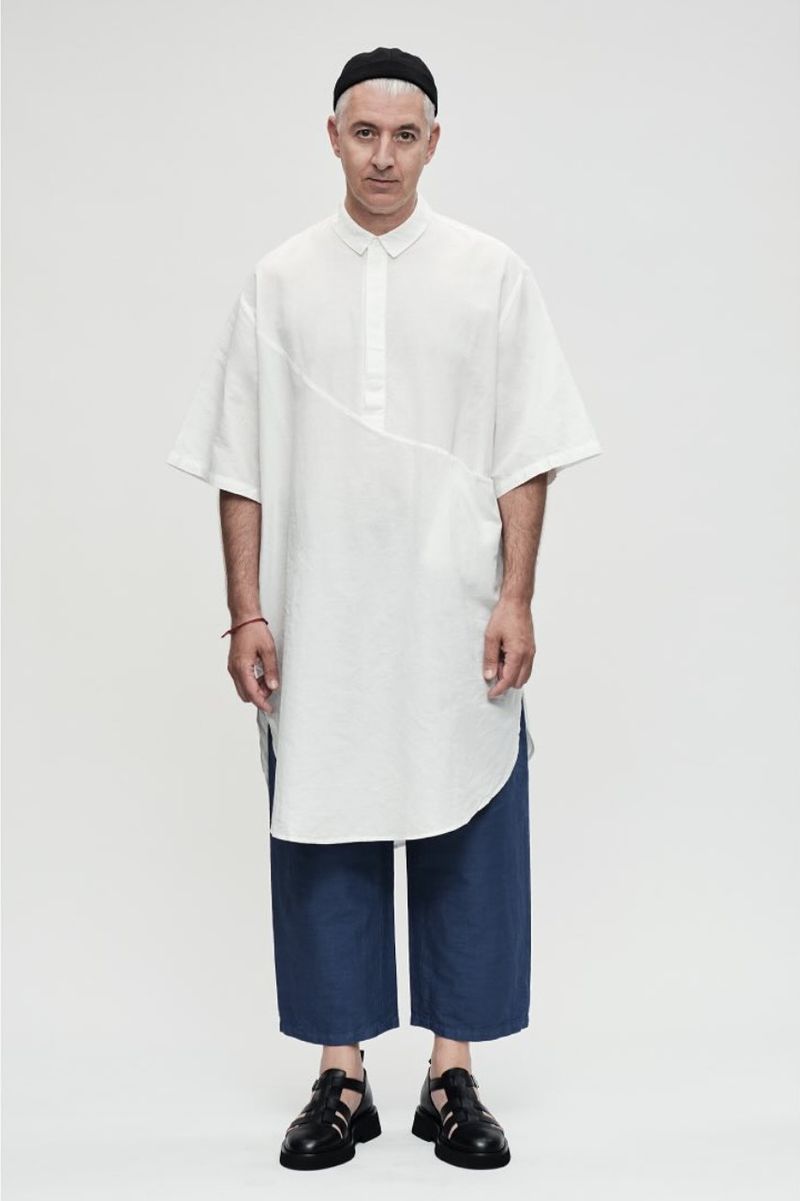Full-length shot of an adult male with short gray hair wearing a white tunic, blue pants, and black sandals against a white background.