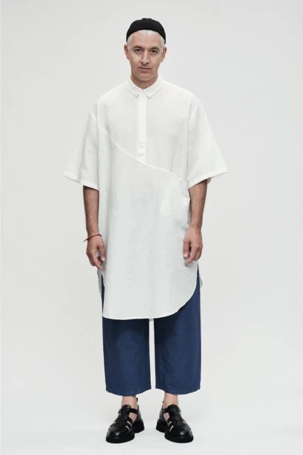Full-length shot of an adult male with short gray hair wearing a white tunic, blue pants, and black sandals against a white background.