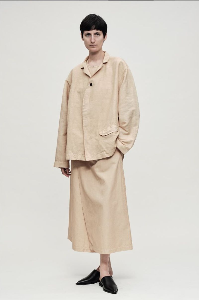 A woman poses in a beige jacket and skirt, paired with black flats against a light background, showcasing minimalist fashion.