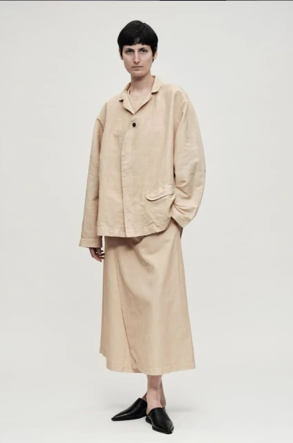 A woman poses in a beige jacket and skirt, paired with black flats against a light background, showcasing minimalist fashion.