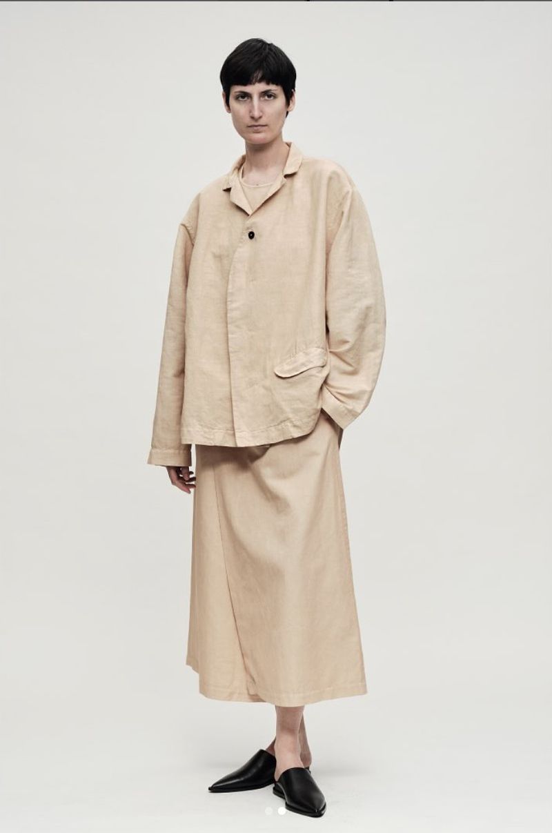 Full-length studio shot of a young woman in a beige suit, with a matching shirt and skirt, paired with black, pointed-toe heeled shoes.