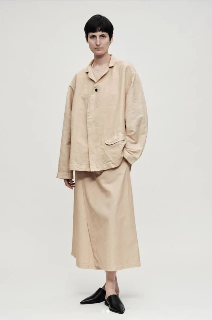 Full-length studio shot of a young woman in a beige suit, with a matching shirt and skirt, paired with black, pointed-toe heeled shoes.