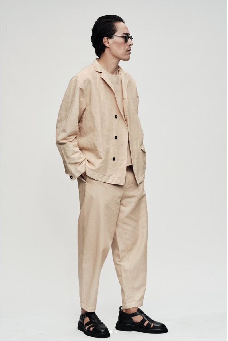 Man in a relaxed beige double-breasted suit, sunglasses, and black leather sandals standing against an off-white studio backdrop.