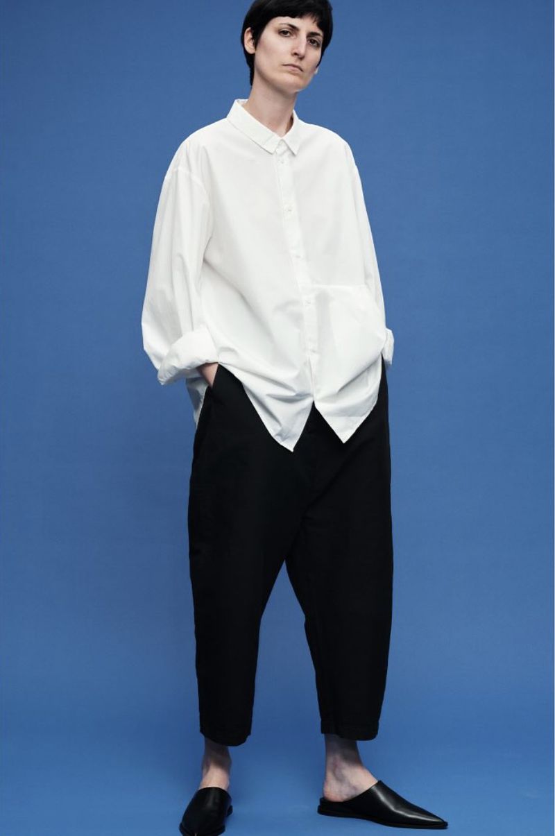 A slender adult fashion model poses in a white, collared, button-down shirt and black trousers, accessorized with matching black leather loafers.
