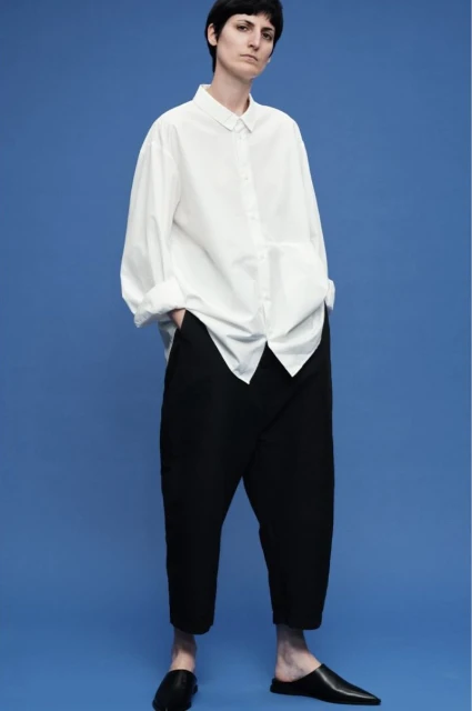A slender adult fashion model poses in a white, collared, button-down shirt and black trousers, accessorized with matching black leather loafers.