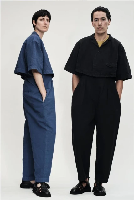 Two models in a photo studio showcase matching blue and black, short-sleeved, wide-leg jumpsuits, posing against a bright white backdrop.