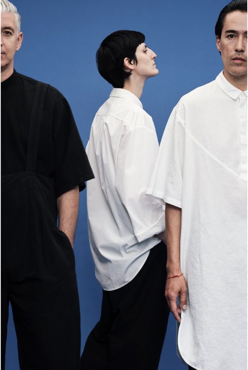 Three models wearing minimalist black and white garments standing against a solid blue backdrop.