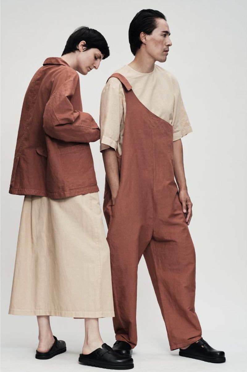 Two models present rust-colored jumpsuits and jackets with neutral skirts against a clean white backdrop, showcasing contemporary fashion.