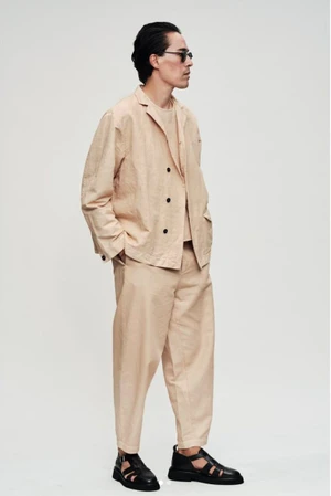 Man in a relaxed beige double-breasted suit, sunglasses, and black leather sandals standing against an off-white studio backdrop.