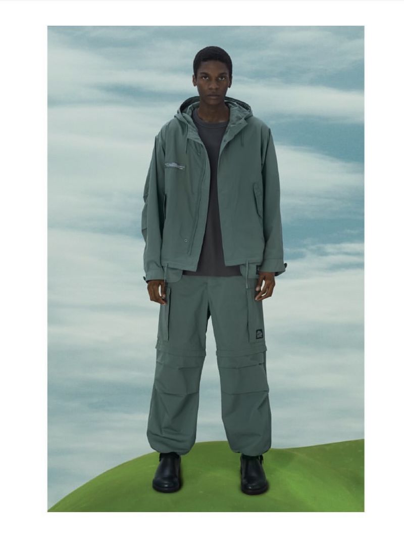 Full-length shot of a Black male model in a hooded jacket and cargo pants against a sky and green hill backdrop in a fashion editorial.