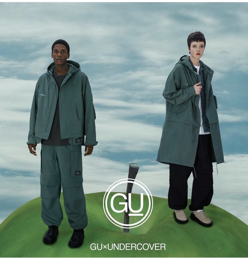 Two fashion models wear green hooded jackets, green pants, black pants and stand on a green apple against a cloudy sky background.