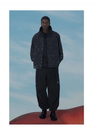 Model Odinaka Ekezie wearing a dark grid pattern overshirt and black cargo pants standing against a painted blue sky backdrop.