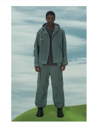 Black man standing outdoors wearing a sage green hooded jacket and matching cargo pants against a sky backdrop
