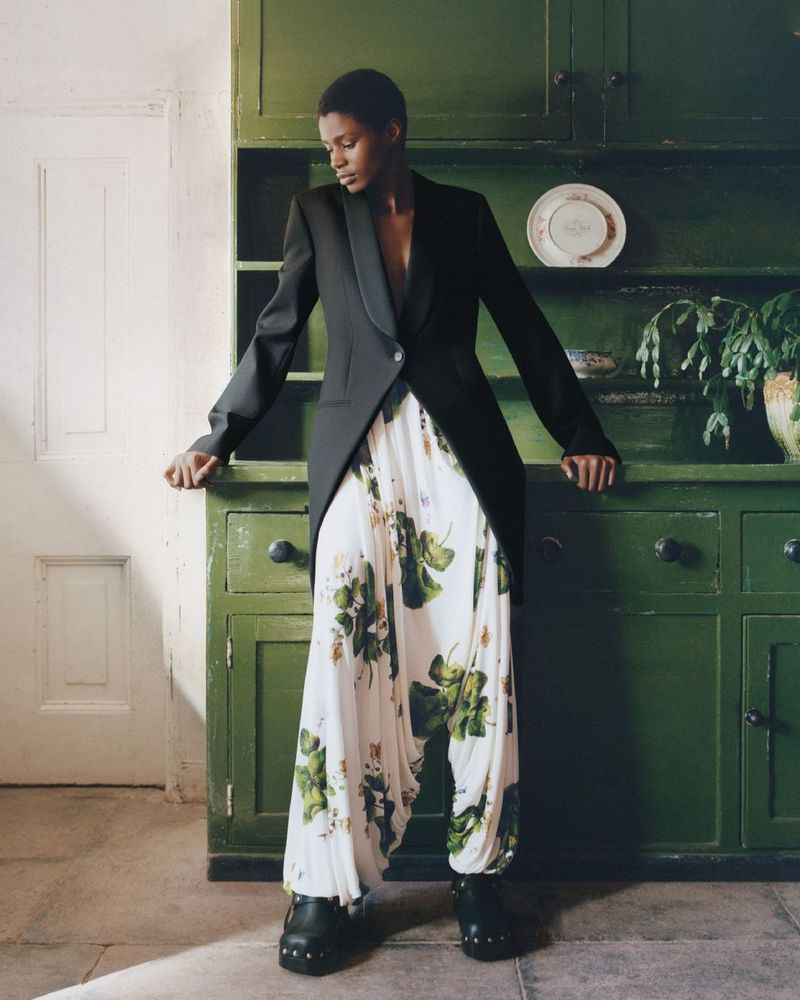 A woman in a black tuxedo jacket and floral print pants leans on a green cupboard in an indoor environment, showcasing fashion and style.