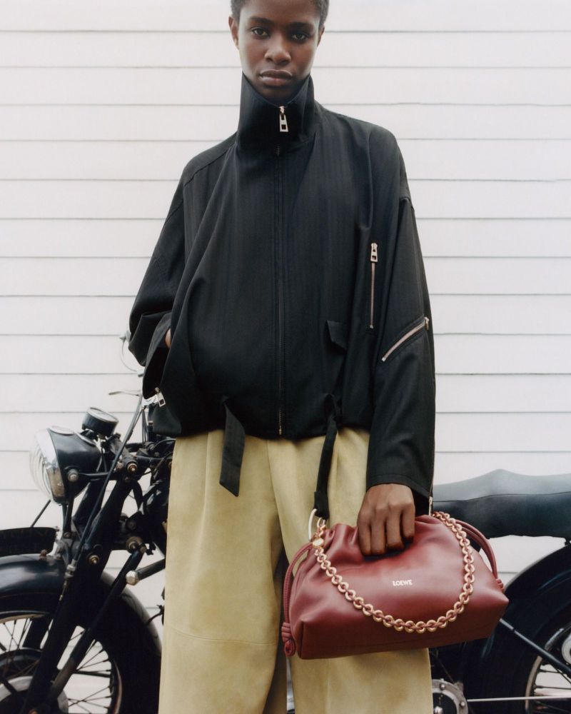 A dark-skinned woman poses with a burgundy LOEWE purse and a motorcycle in the background, wearing a black bomber jacket and khaki pants.
