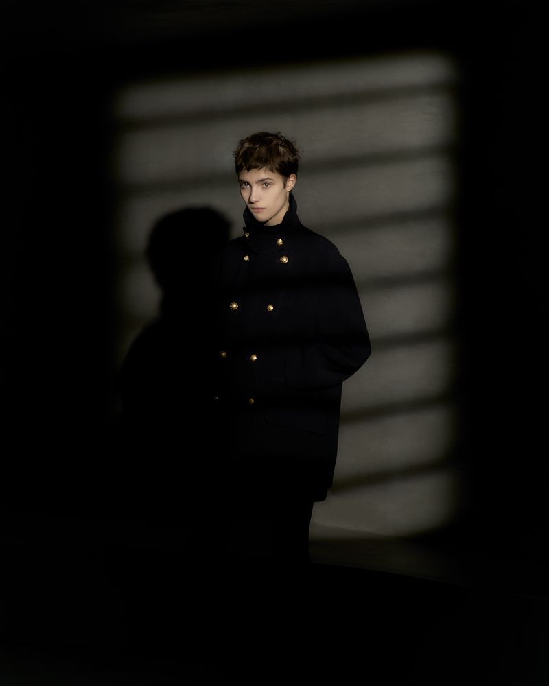 Portrait of a young adult woman with short brown hair wearing a black coat with gold buttons against a dark shadow patterned background.