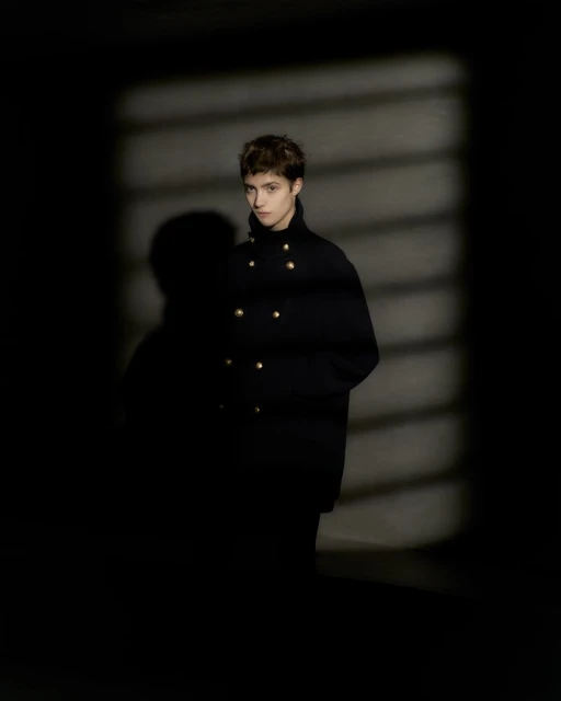 Portrait of a young adult woman with short brown hair wearing a black coat with gold buttons against a dark shadow patterned background.