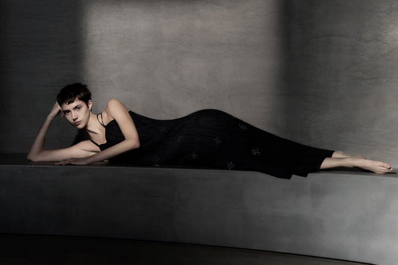 A young adult female model with short brown hair in a black dress lies on a grey surface indoors in a fashion photography setting.