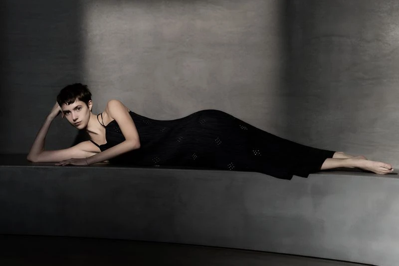 A young adult female model with short brown hair in a black dress lies on a grey surface indoors in a fashion photography setting.