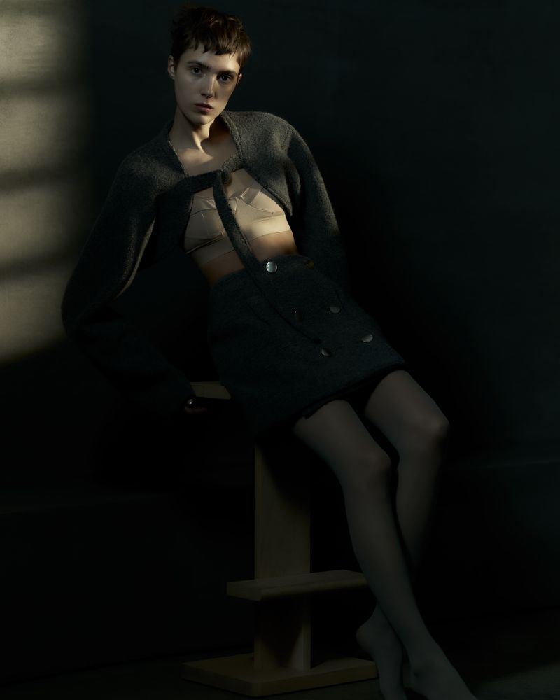 Fashion shot of a slender young woman in a cardigan and skirt, seated on a wooden stool in soft, muted lighting against a dark backdrop.