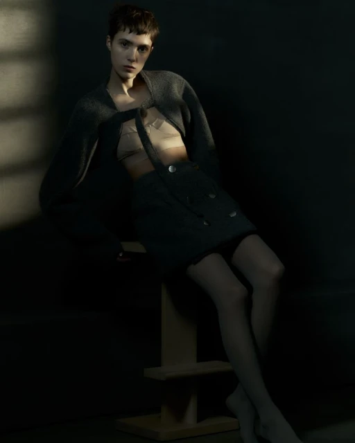 Fashion shot of a slender young woman in a cardigan and skirt, seated on a wooden stool in soft, muted lighting against a dark backdrop.