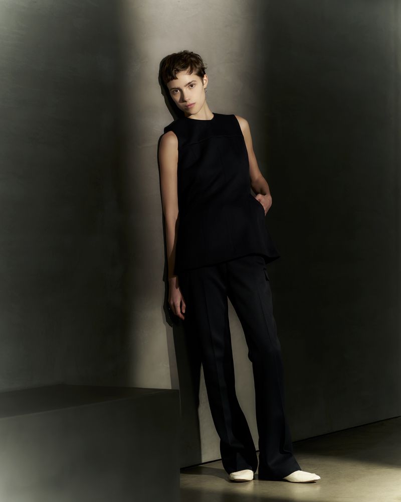 Fashion shot of woman with short hair leaning against a wall wearing a black top and pants in a photo studio with soft lighting.