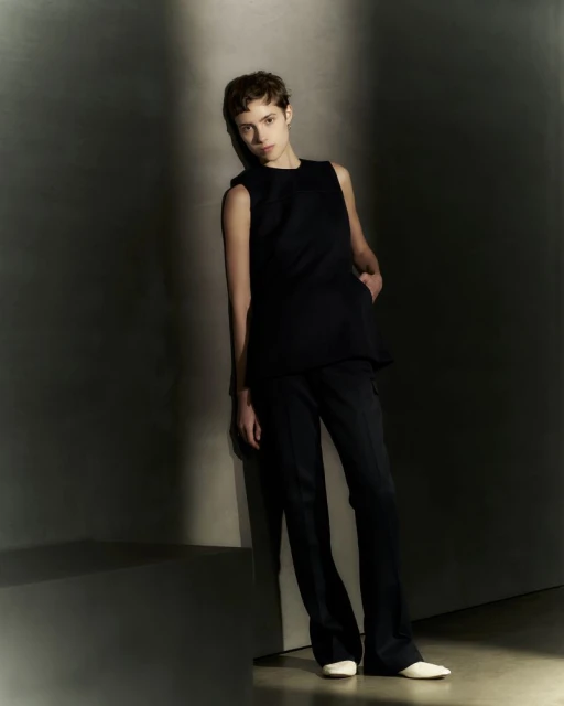 Fashion shot of woman with short hair leaning against a wall wearing a black top and pants in a photo studio with soft lighting.