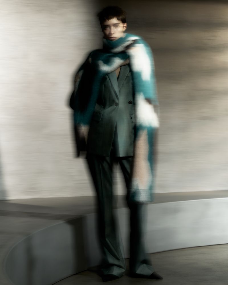 A blurred, full-length shot captures a slender adult in a green suit and patterned scarf, posing indoors with neutral wall background.