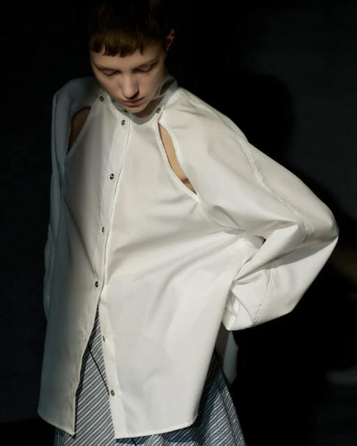 Young woman in cut-out white shirt and striped skirt poses against a dark backdrop in a fashion editorial-style studio portrait.