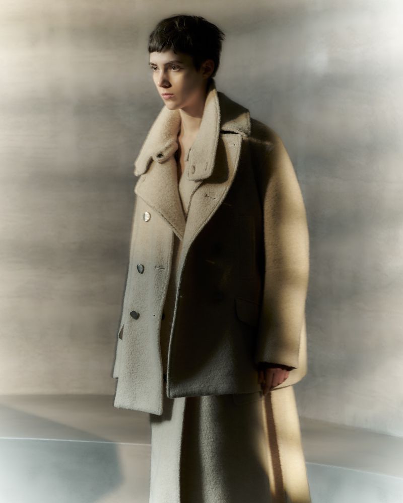 Studio shot of a fair-skinned woman wearing a beige coat and dress, standing in a photo studio with soft, neutral lighting.