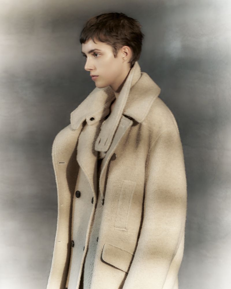 Fashion portrait of a young adult with short brown hair wearing a cream-colored overcoat in front of a neutral gray backdrop.
