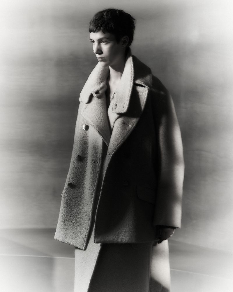 Monochrome portrait of a young adult woman with short dark hair in a wool overcoat and dress, in soft studio lighting against a grey backdrop.