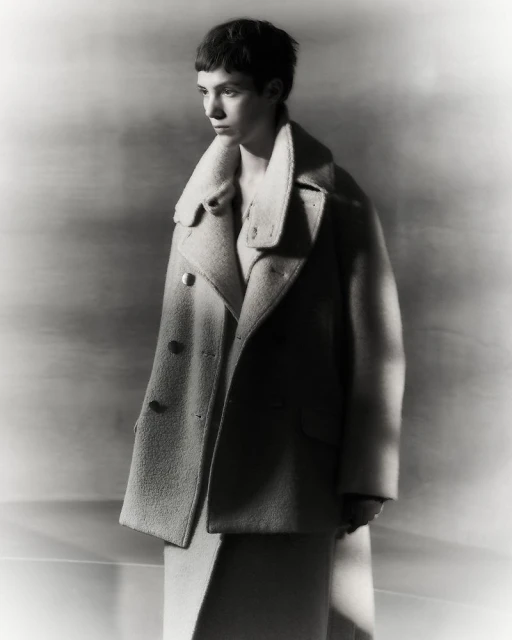 Monochrome portrait of a young adult woman with short dark hair in a wool overcoat and dress, in soft studio lighting against a grey backdrop.