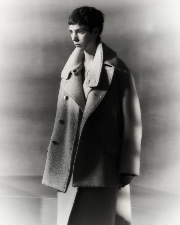 Monochrome portrait of a young adult woman with short dark hair in a wool overcoat and dress, in soft studio lighting against a grey backdrop.