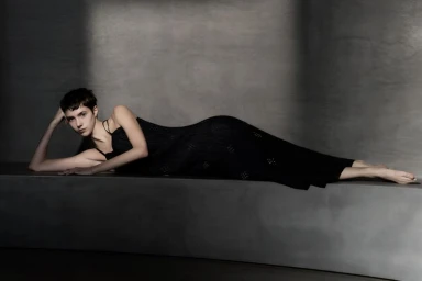 A woman with short dark hair in a black slip dress reclines on a grey concrete ledge against a textured grey wall.