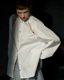 Model looking down wearing an oversized white button-up shirt with shoulder cutouts against a dark studio background.