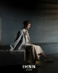 Model with short brown hair seated on a curved grey platform wearing an oversized jacket and cream skirt for IHNN.