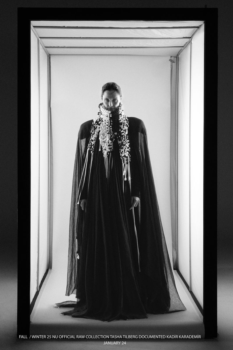 Monochrome fashion shot of a person in a black gown and decorative collar, framed by a rectangular light installation in a studio setting.