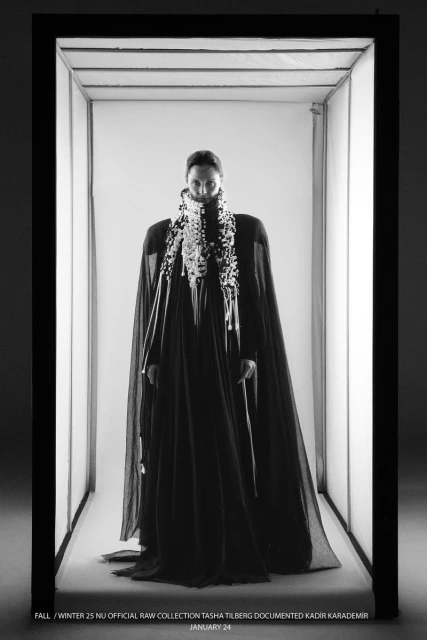 Monochrome fashion shot of a person in a black gown and decorative collar, framed by a rectangular light installation in a studio setting.