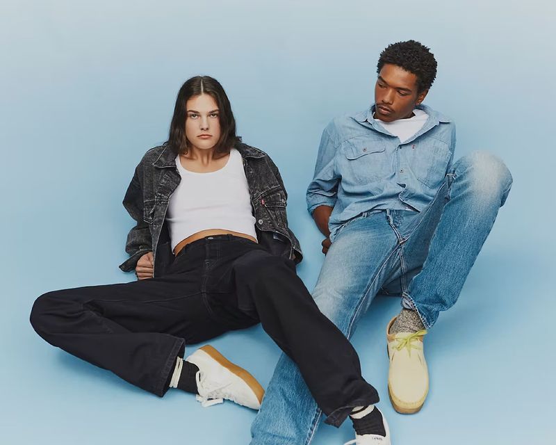 Two people, an adult woman and an adult male, are sitting indoors against a light blue backdrop wearing denim and sneakers, for a fashion shot.