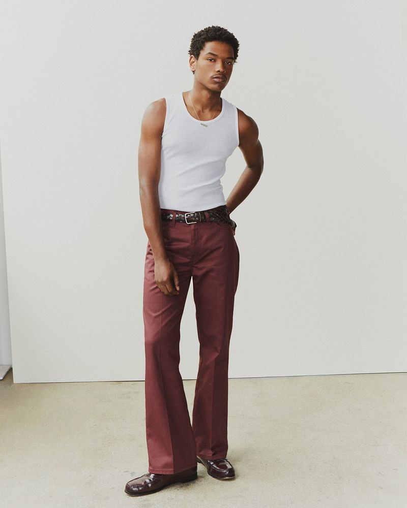 Full-body portrait of a Black male model wearing a white tank top and maroon bell-bottom pants, posing in a photo studio.