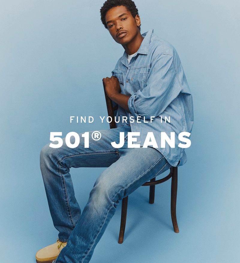 Man sits in light blue denim shirt and jeans, leaning back on a chair in a photo studio with text promoting 501 jeans.
