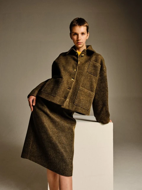A young woman poses in a studio wearing a dark green jacket and skirt, complemented by a yellow top, exuding style and confidence.
