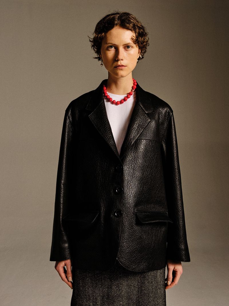 Woman in a black, textured blazer and red beaded necklace poses indoors. Neutral background, medium shot, fashion editorial.