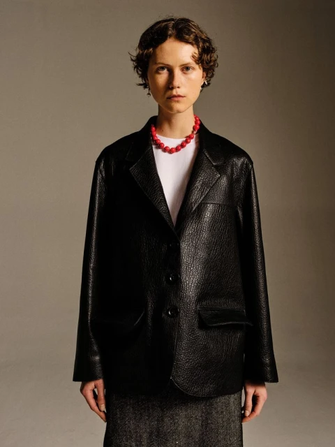 Woman in a black, textured blazer and red beaded necklace poses indoors. Neutral background, medium shot, fashion editorial.