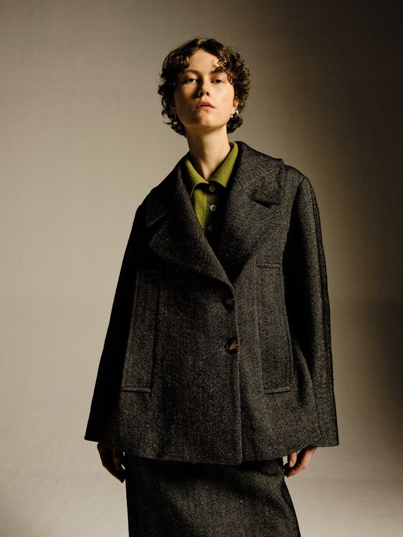 Portrait of a woman with short curly hair wearing a grey tweed blazer and matching skirt, styled in a photo studio setting.