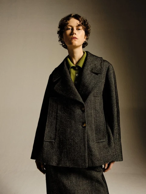 Portrait of a woman with short curly hair wearing a grey tweed blazer and matching skirt, styled in a photo studio setting.