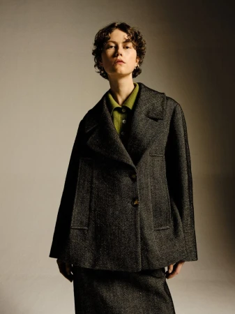Portrait of a woman with short curly hair wearing a grey tweed blazer and matching skirt, styled in a photo studio setting.
