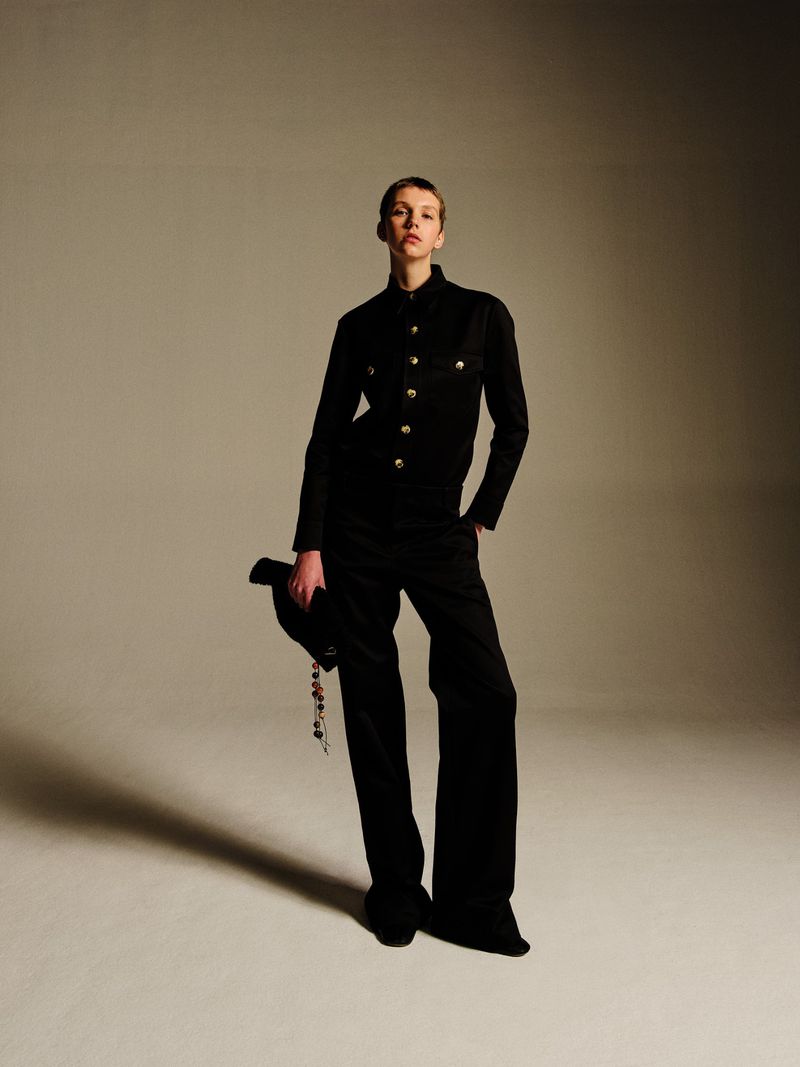 A fair-skinned woman with short hair poses in a black jacket and wide-legged pants, holding a hat indoors against a neutral backdrop.