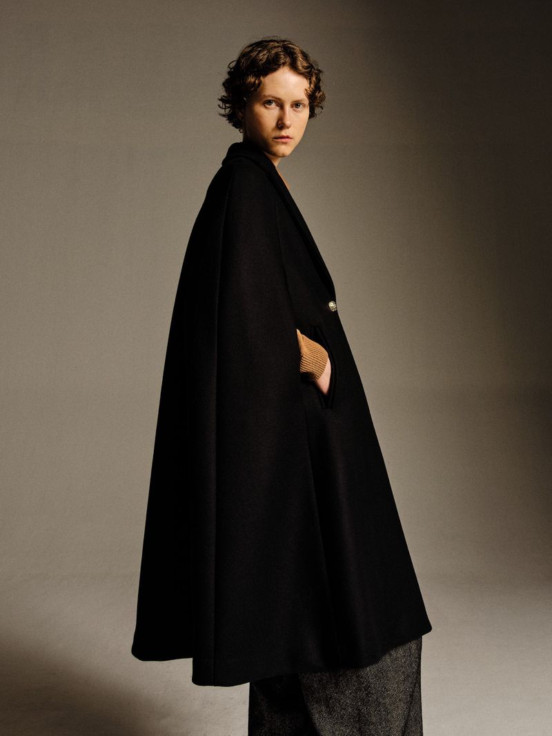 Studio shot of a stylish woman wearing a black cape and grey trousers, posing against a neutral grey background, demonstrating fashion apparel.